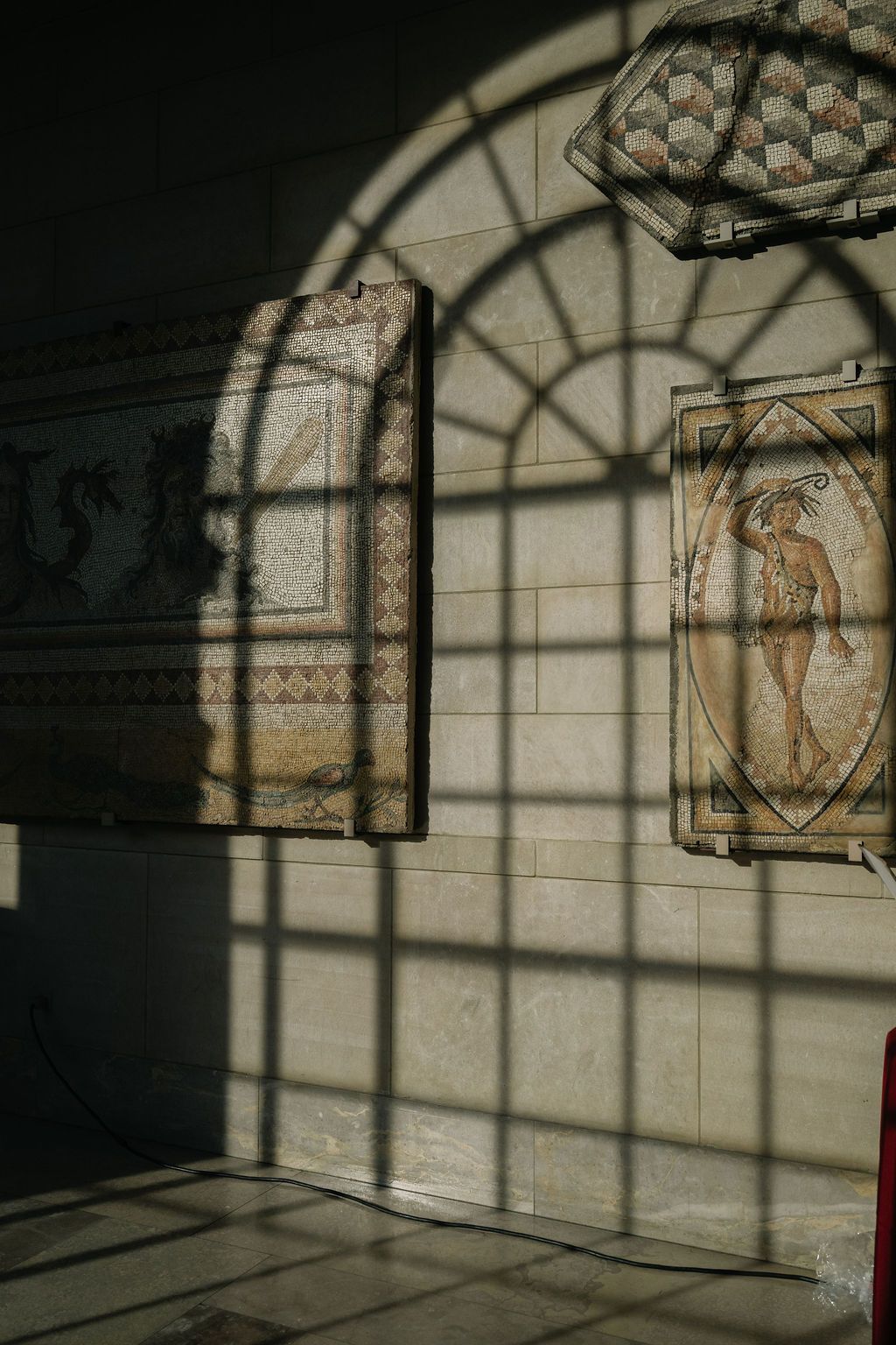 Sunlight casts geometric shadows through an arched window onto a stone wall with ornate decorative panels.