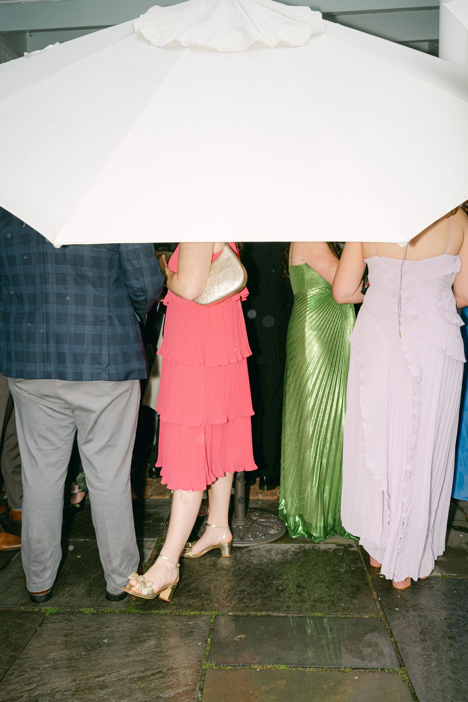 Group of wedding guests in colorful formal attire sheltering under a large white patio umbrella on a rainy day.