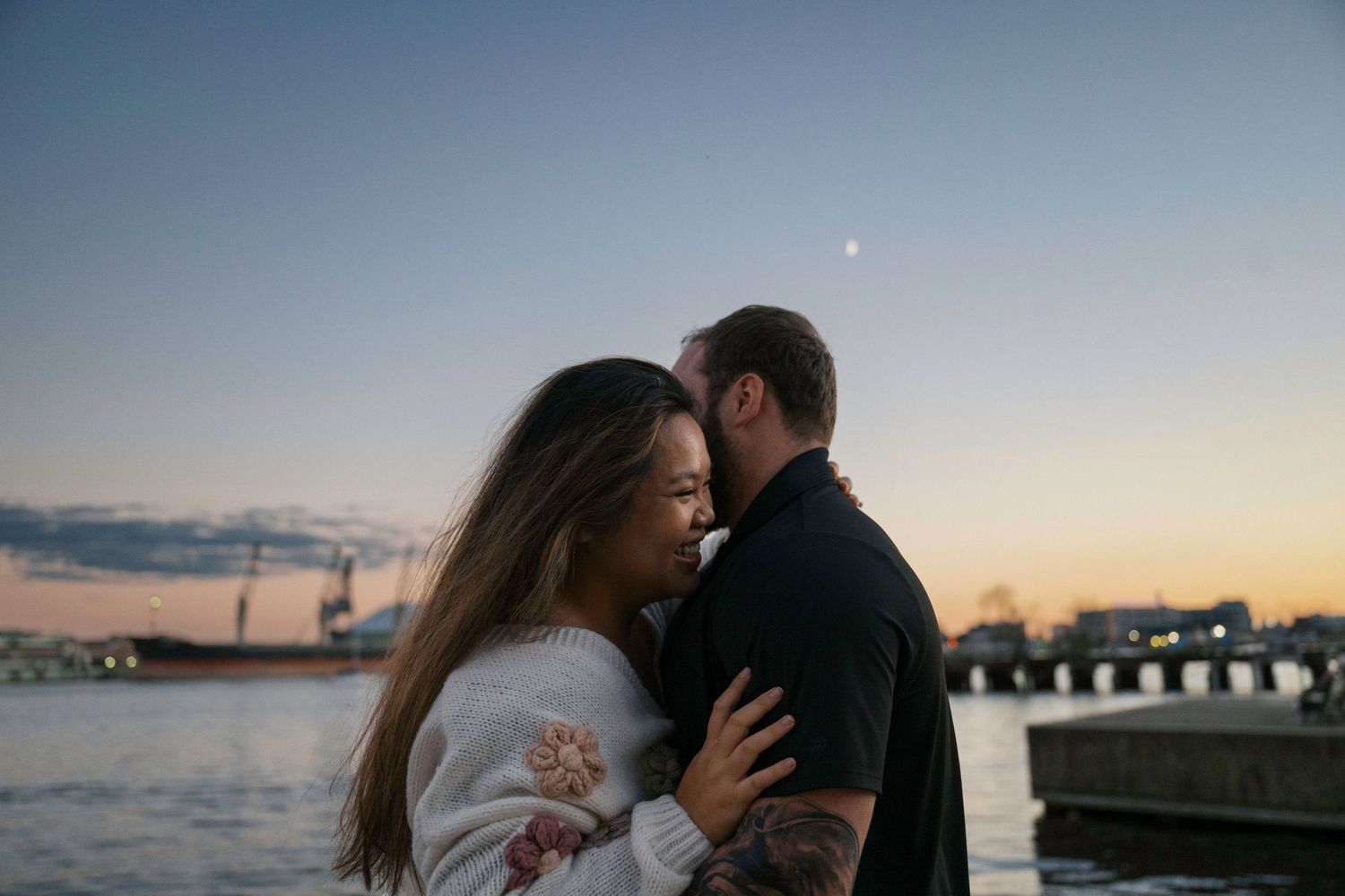 Romantic couple embraces at sunset by the waterfront with a pier and glowing sky in the background.