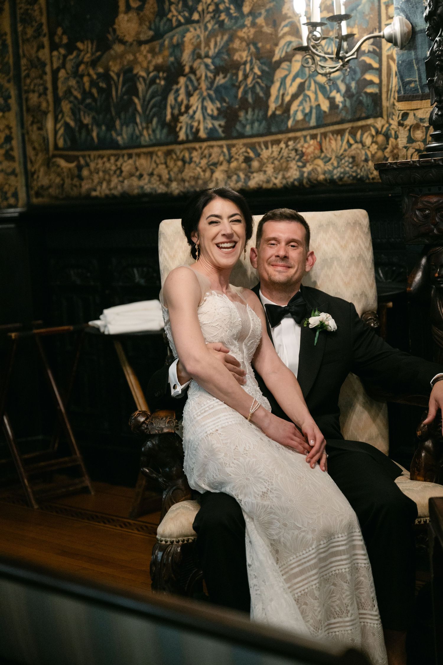 Newlyweds share a joyful moment on an ornate sofa against a vintage wallpapered wall at their wedding reception.