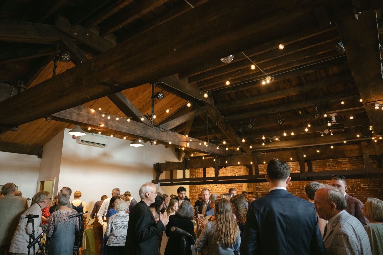 A large group gathers under rustic wooden beams and string lights at an indoor venue during an event.