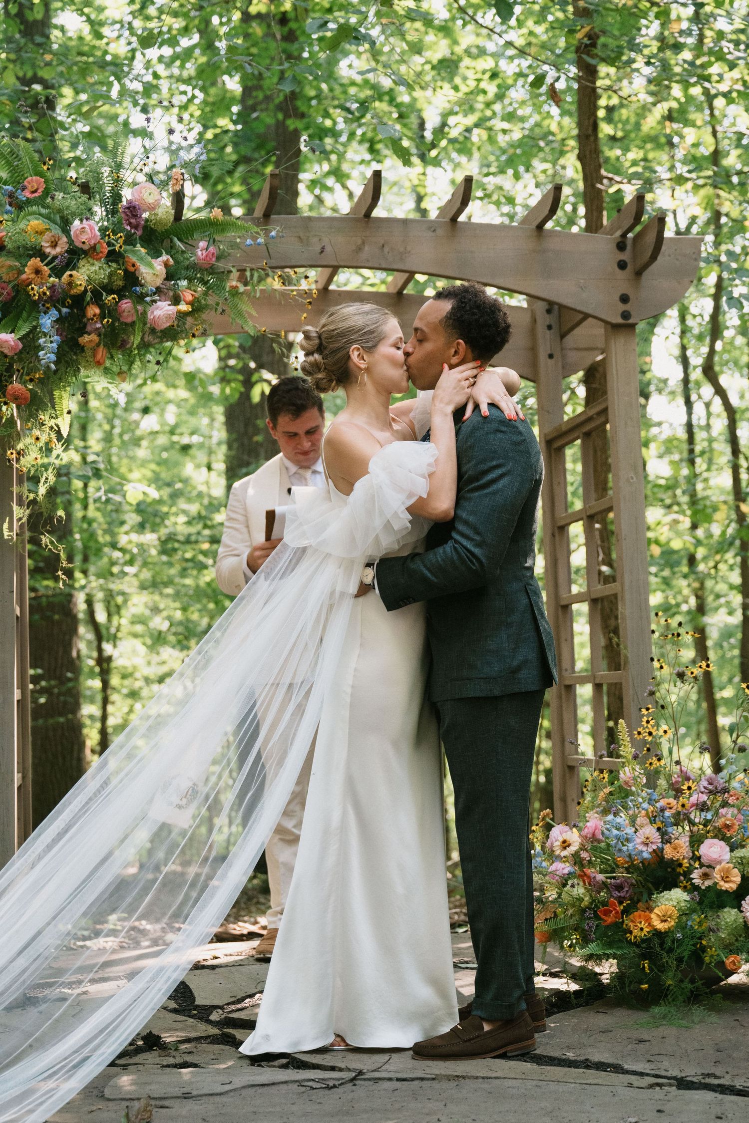 Newlyweds share their first kiss under a flower-decorated wooden arch in a forest.