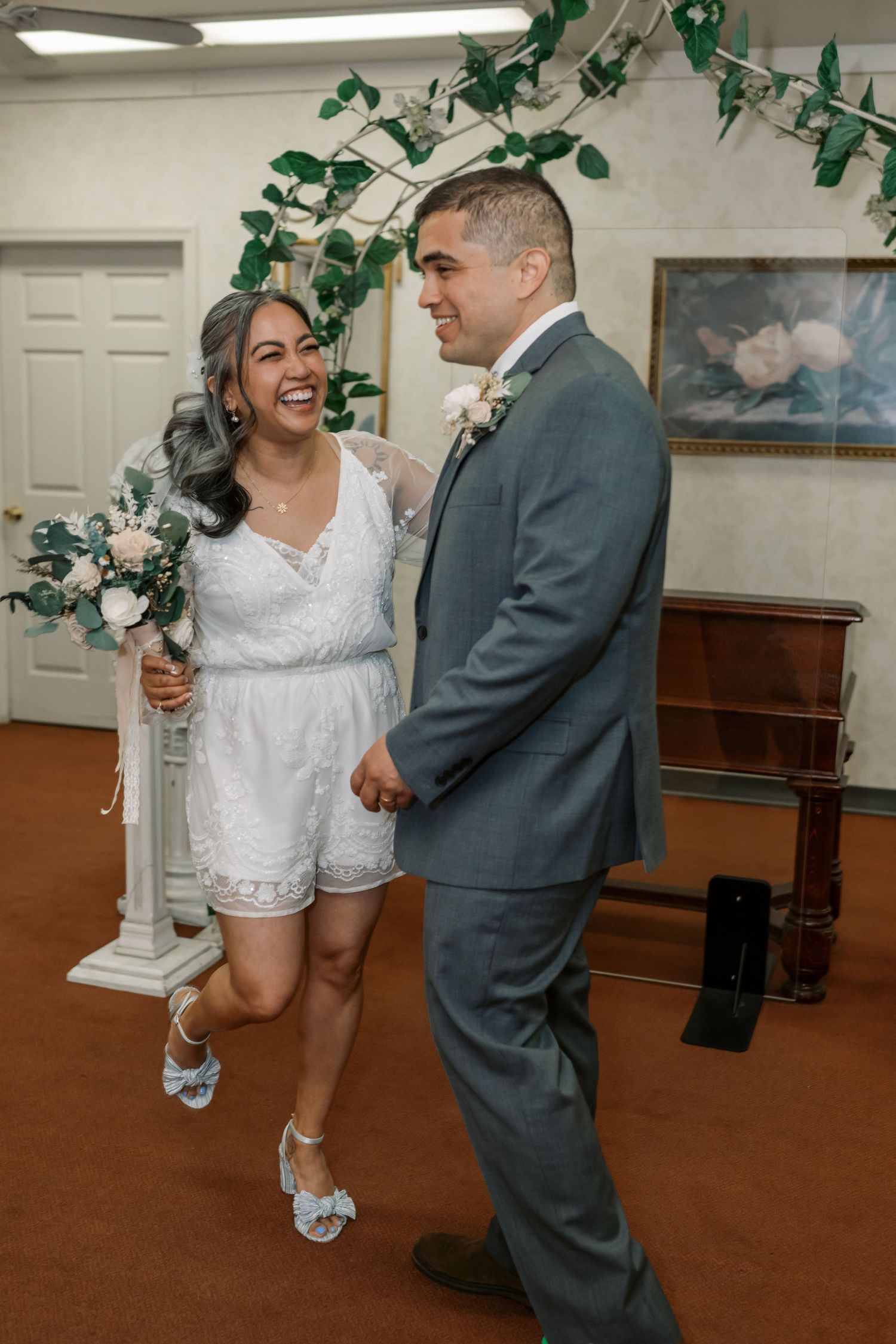 A wedding couple shares a joyful moment under green foliage decorations in a carpeted indoor ceremony space.
