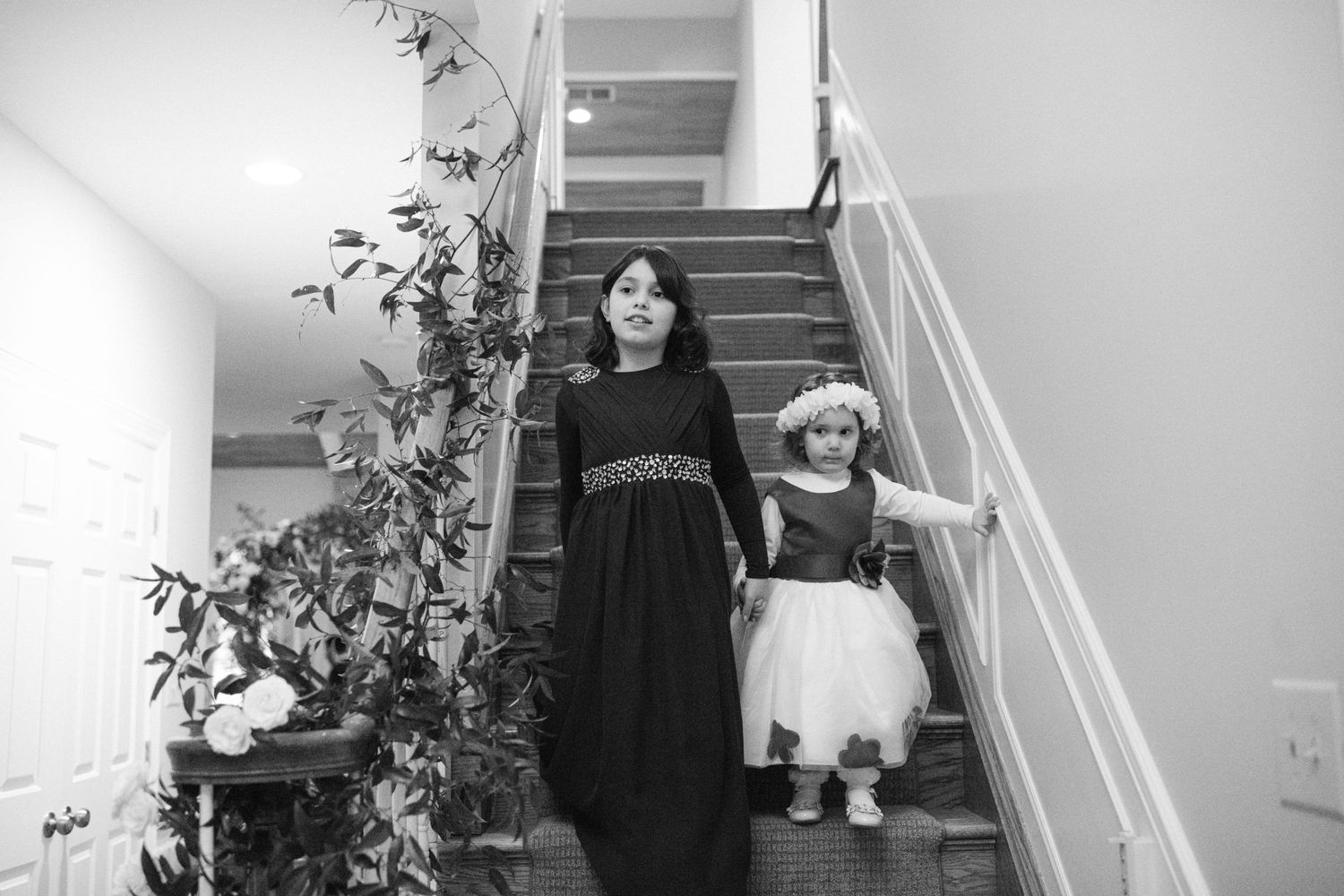 Two figures stand on a staircase with a houseplant against a white wall, captured in black and white.