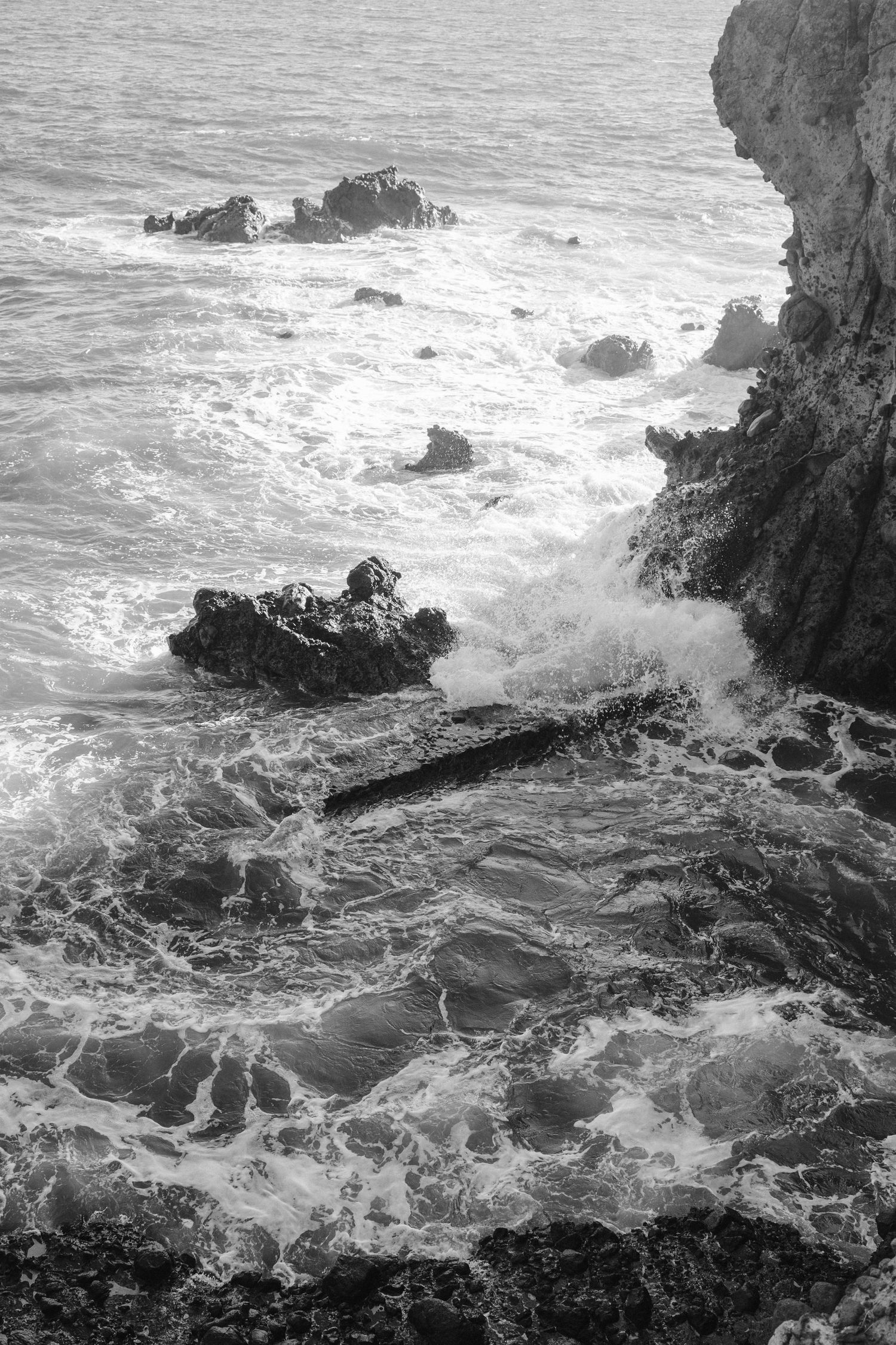 Dramatic black and white seascape showing waves crashing against rocky cliffs and outcrops along a rugged coastline.