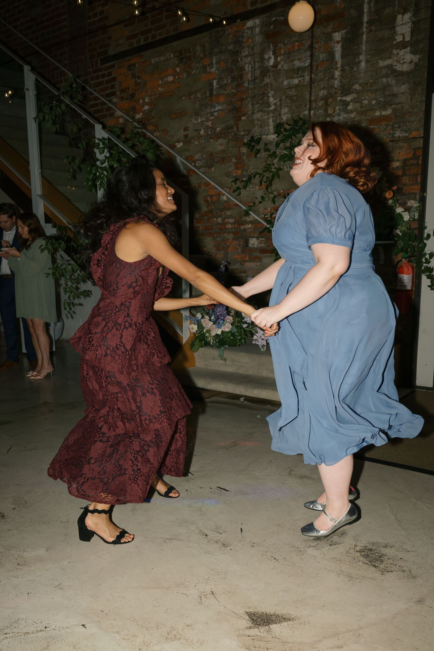 Two dancers in elegant dresses holding hands and dancing enthusiastically on a concrete floor.