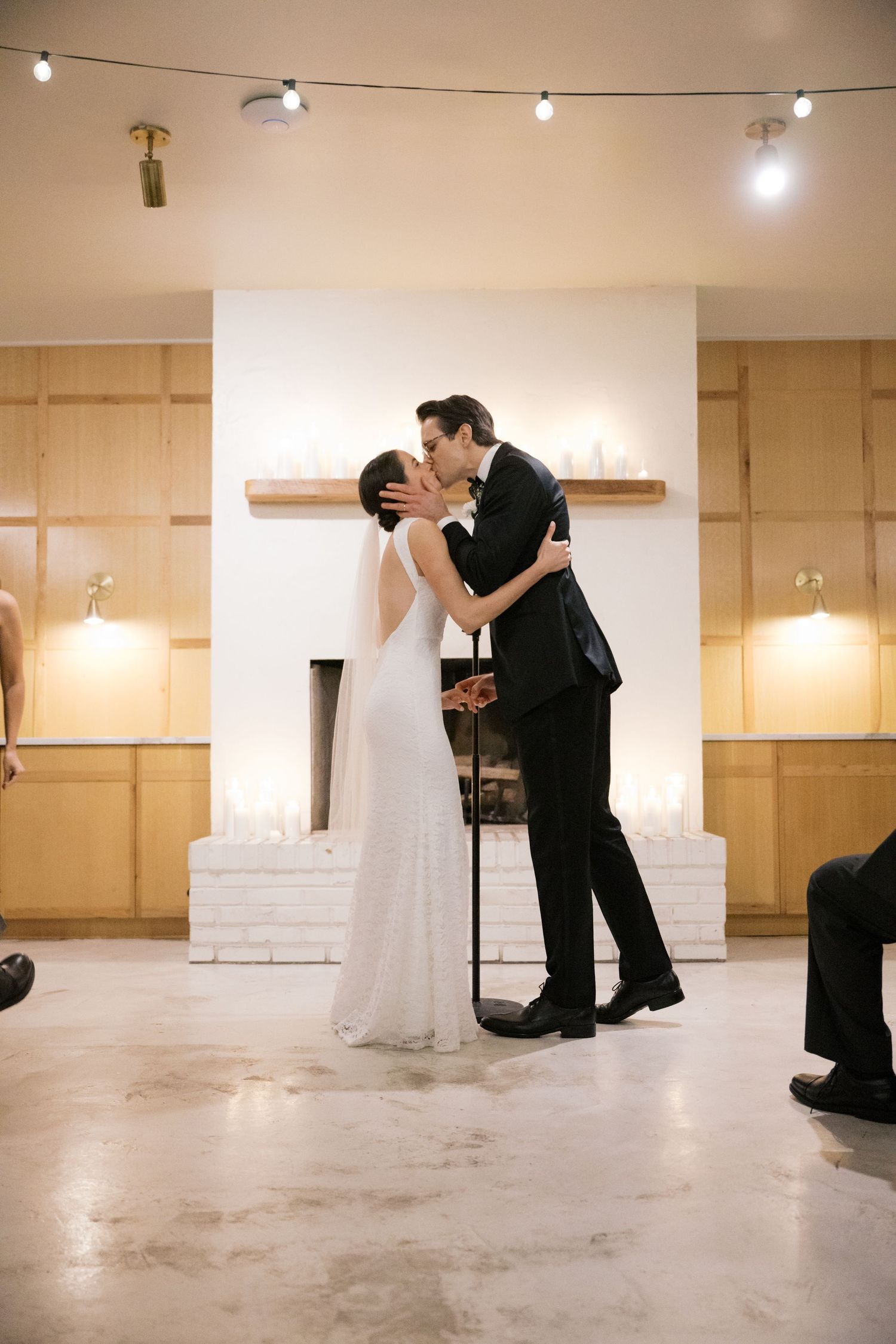 A romantic wedding ceremony moment with a couple kissing in front of a white brick fireplace under string lights.