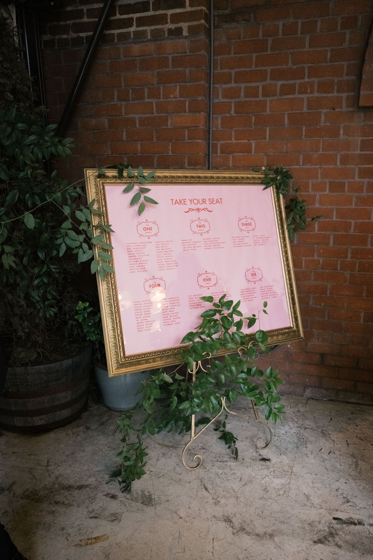A pink wedding seating chart in an ornate gold frame decorated with greenery against a brick wall.