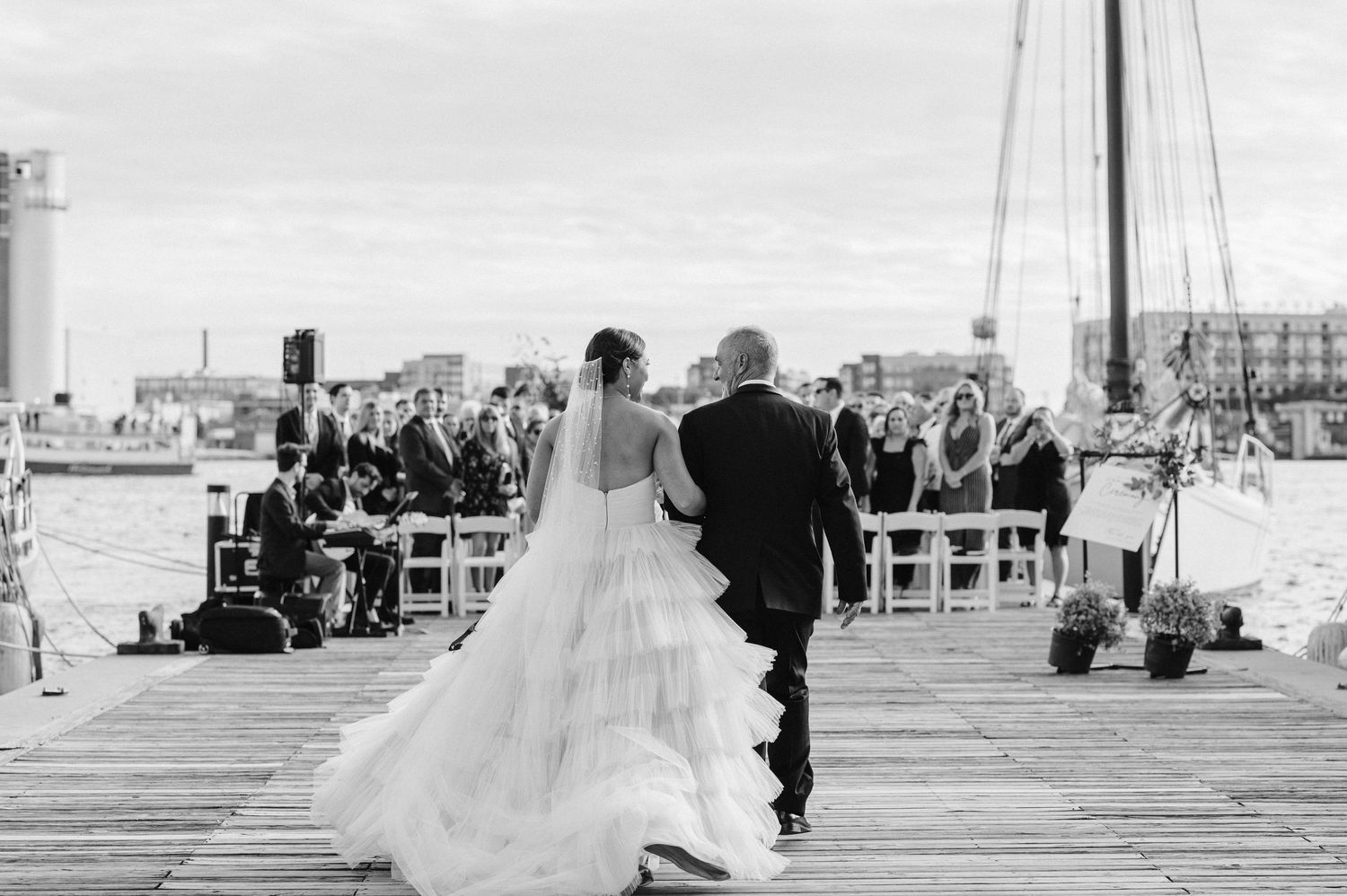 Black and white photo of wedding ceremony on waterfront pier with seated guests.