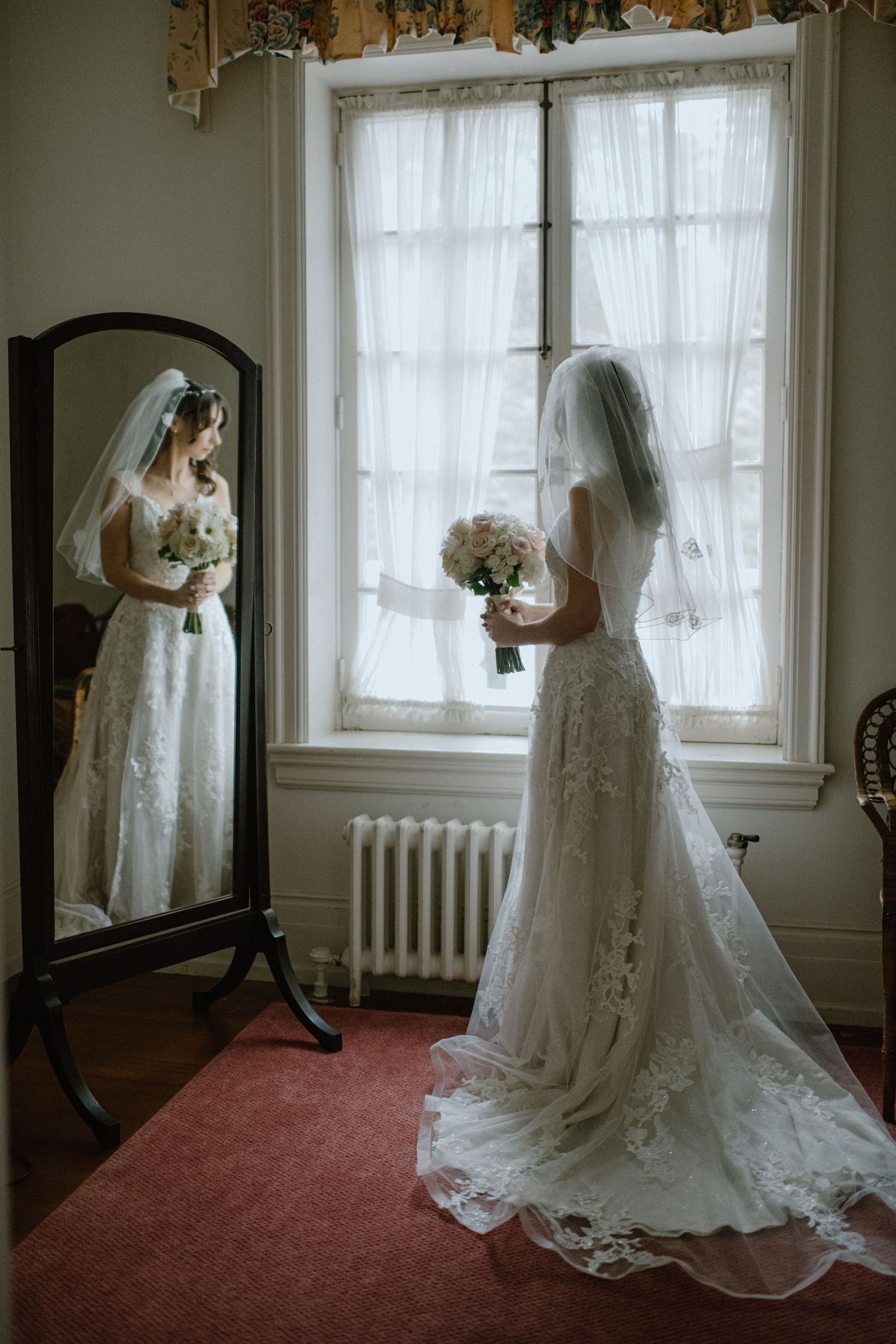 Bride in lace wedding gown looking at her reflection in a vintage mirror by a window.