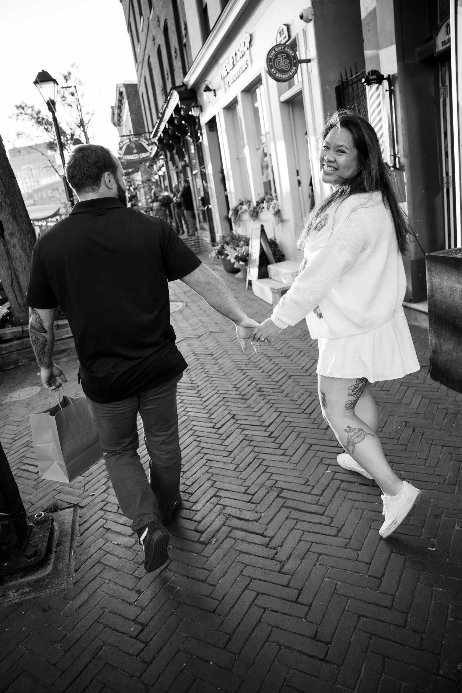 A joyful monochrome candid shot of two people dancing together on a brick sidewalk in front of storefronts.