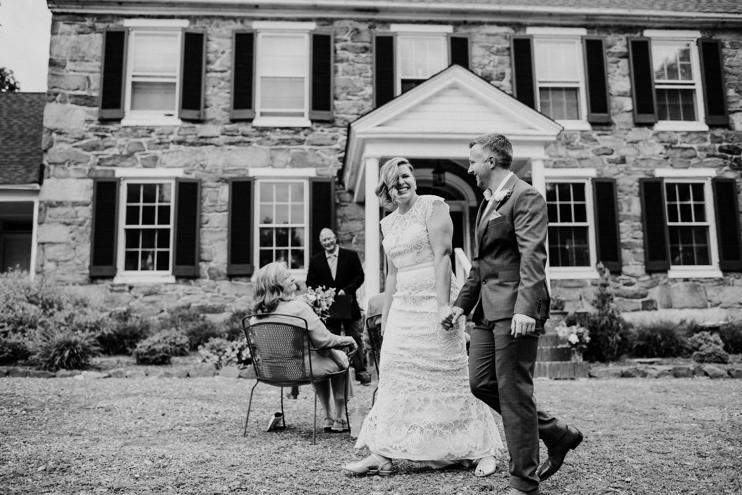 A black and white wedding photo in front of a historic stone mansion with white shuttered windows.