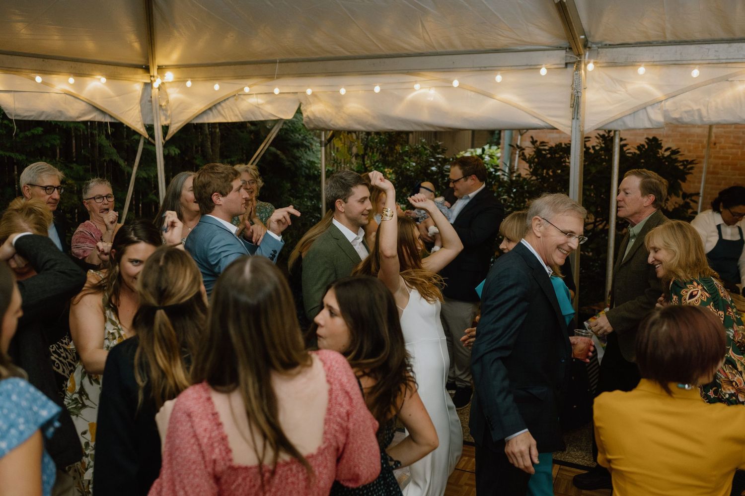 Wedding guests dance and celebrate under a white tent with string lights during an outdoor reception.