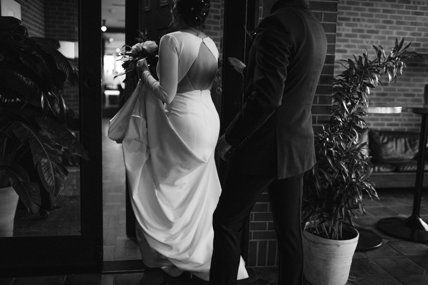 Black and white photo of a couple in wedding attire standing near a potted plant.
