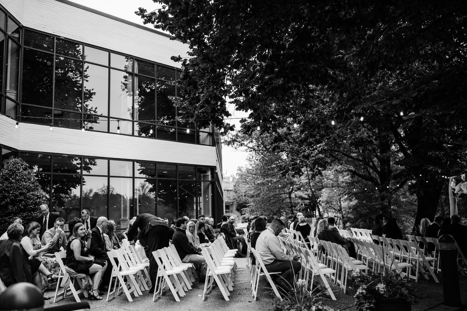 Black and white photo of wedding guests seated in rows of white chairs during outdoor ceremony.