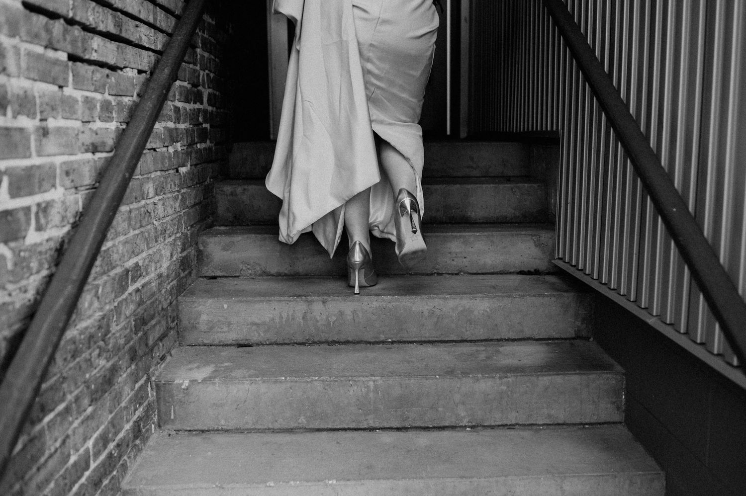 A flowing wedding dress and feet descending concrete stairs in a moody black and white photograph.