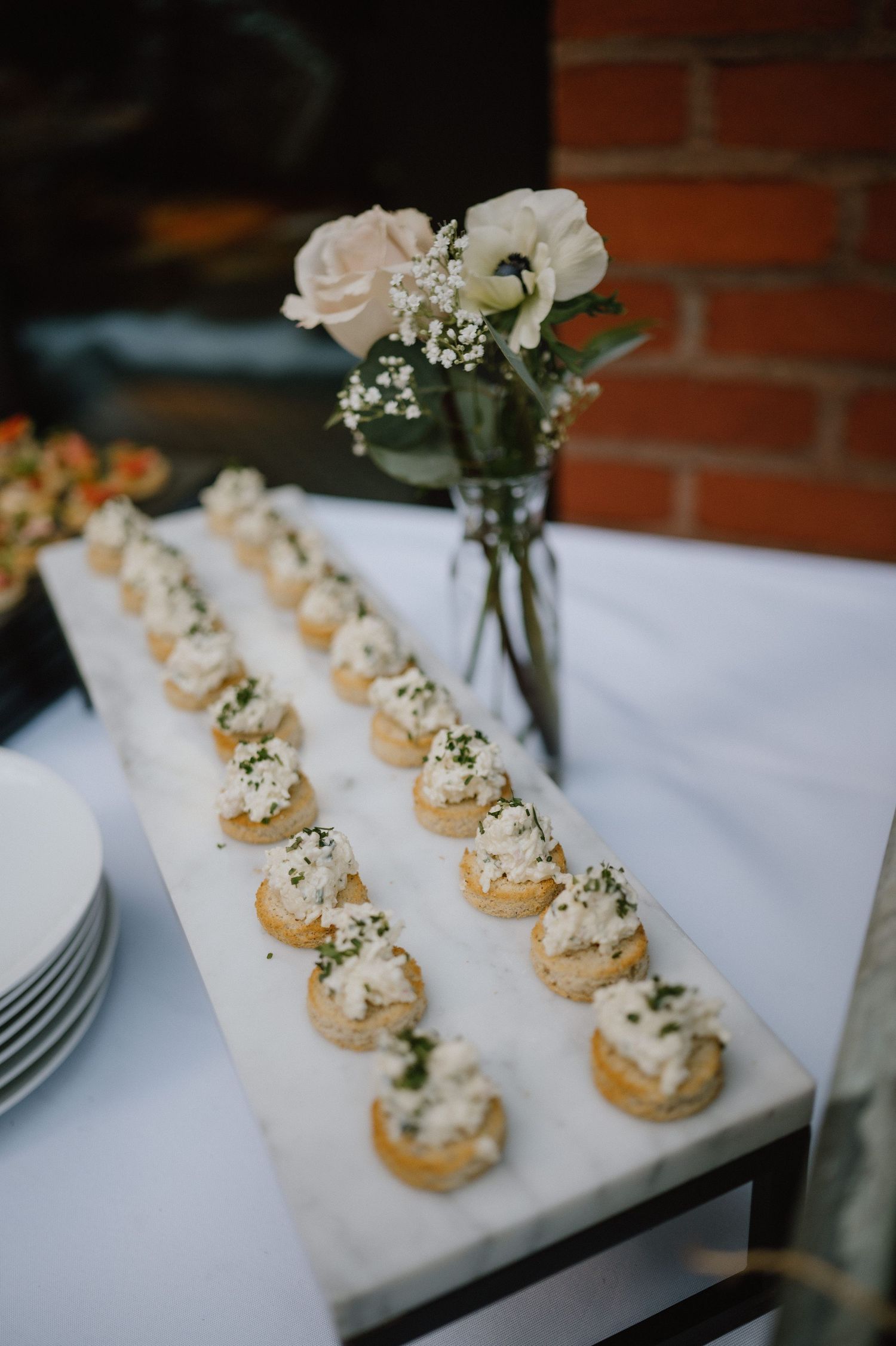Elegant appetizers arranged in neat rows on a white tablecloth next to a vase of pale pink roses and baby's breath.