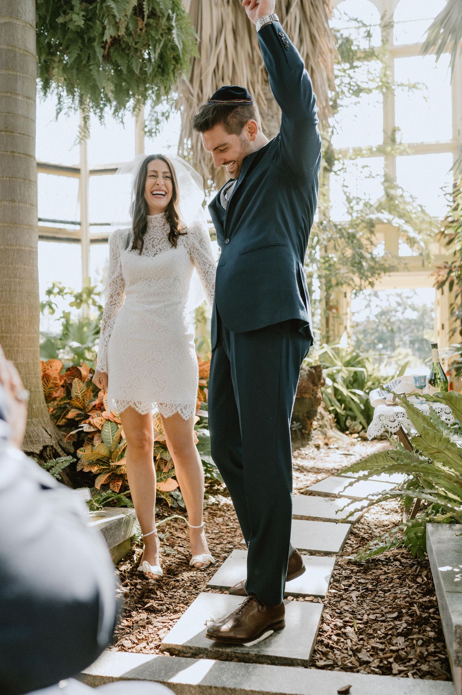 A celebratory moment in a greenhouse with a short white lace dress and navy suit during an intimate ceremony.