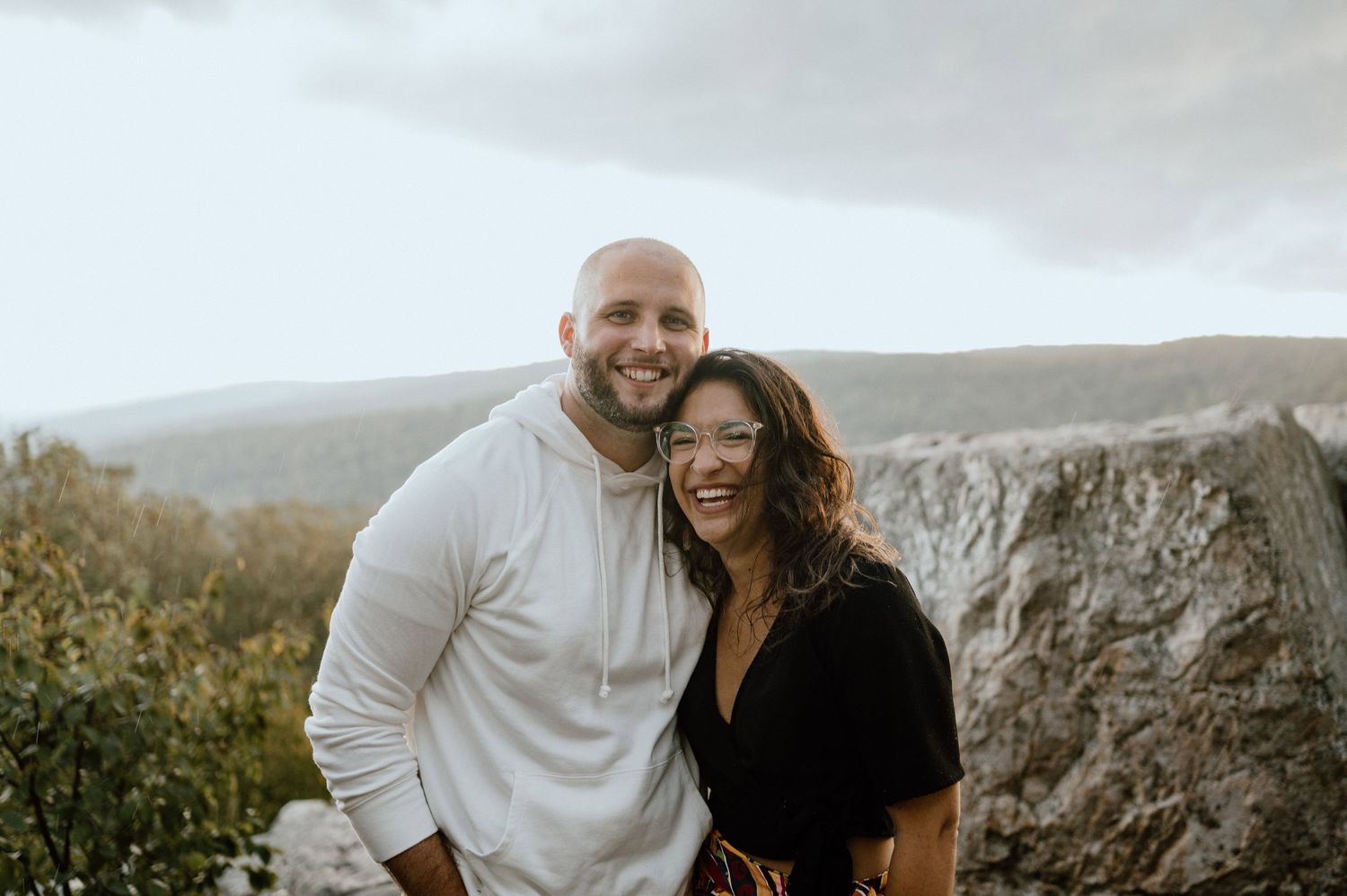 A romantic couple smile together while standing on a mountain overlook with dramatic rocky cliffs behind them.