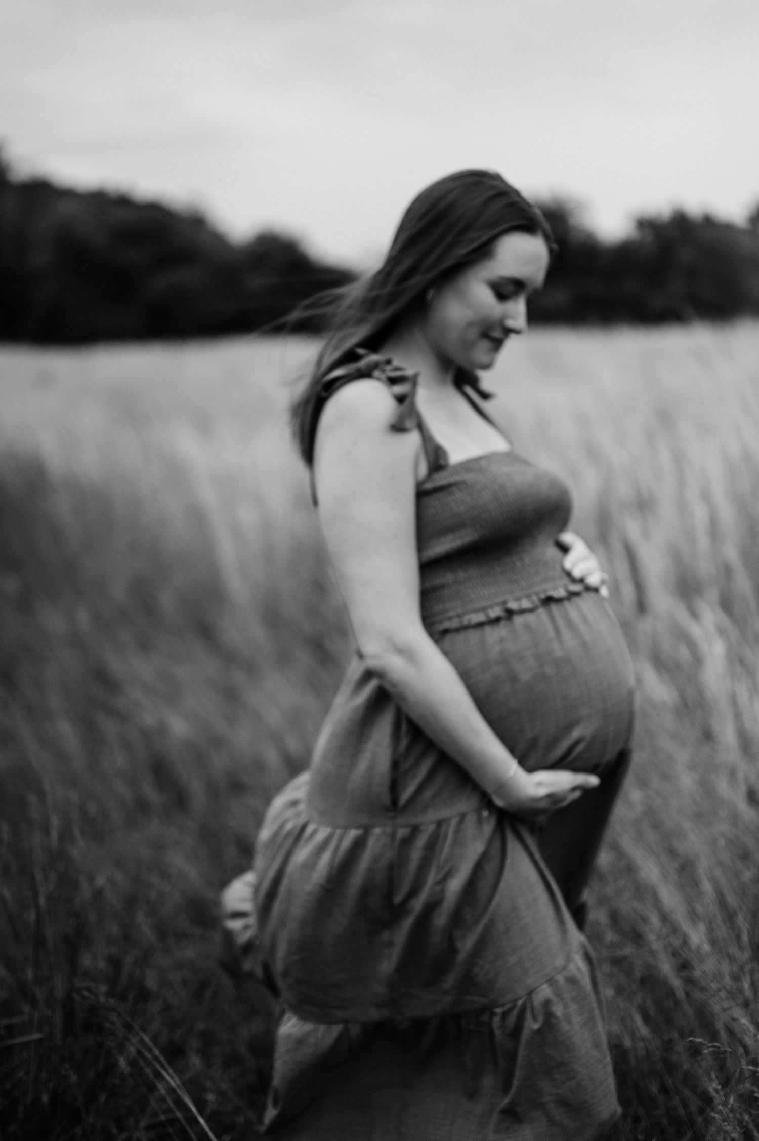 A black and white portrait of an expectant mother in a flowing dress standing in tall grass.