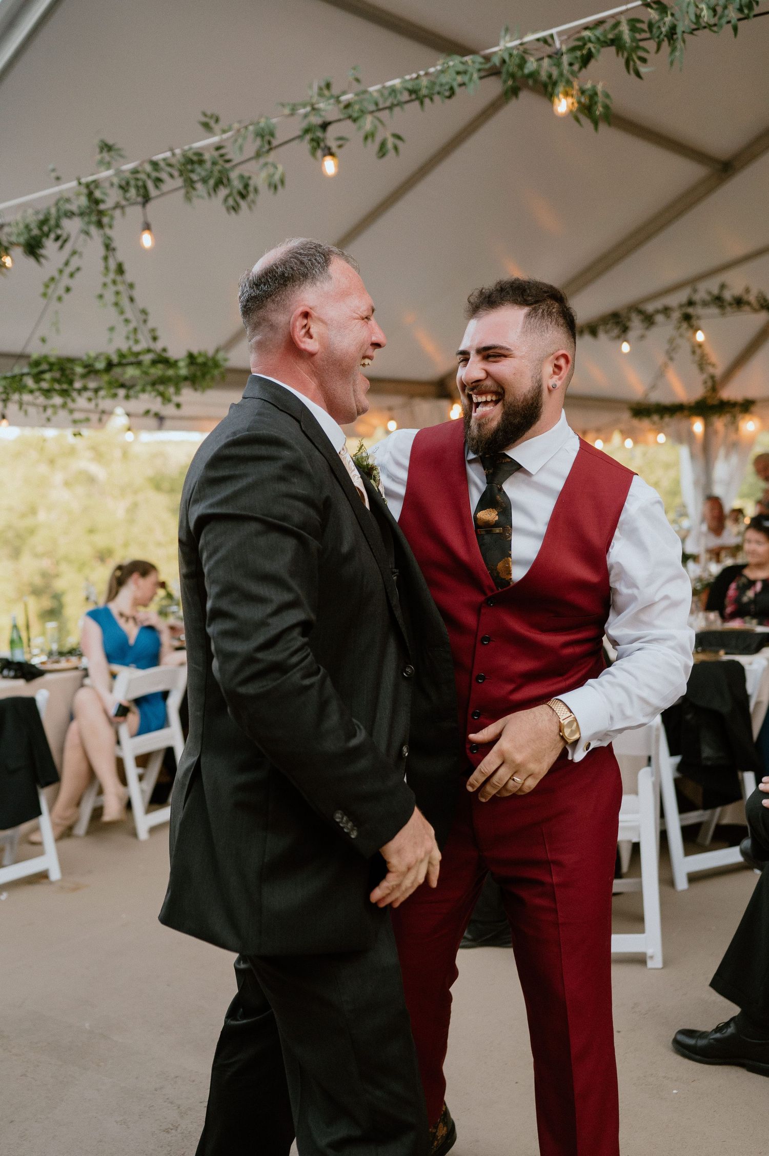 Two wedding guests share a lighthearted moment while dancing under a tent with string lights.