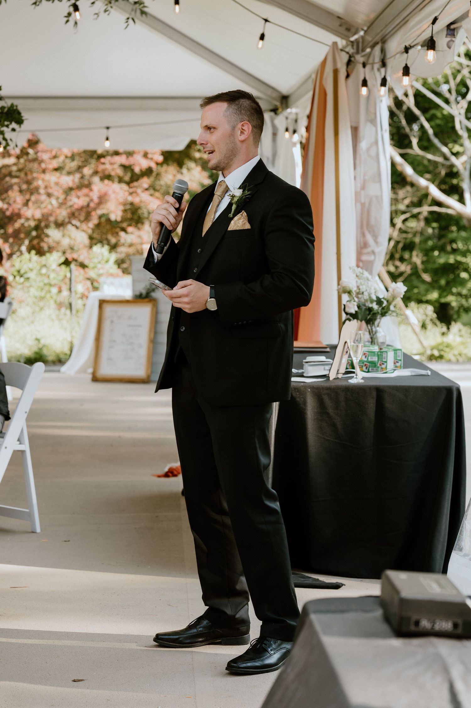 Wedding guest delivers heartfelt speech during outdoor reception under white tent with string lights.