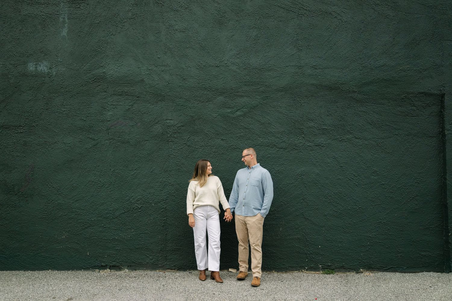Couple holding hands standing against a dark green wall outdoors, smiling at each other in casual attire.