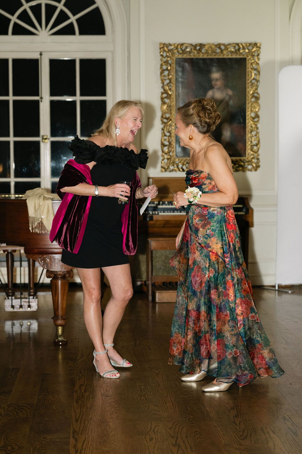 Two women dancing and laughing together at an elegant indoor reception with ornate decor.