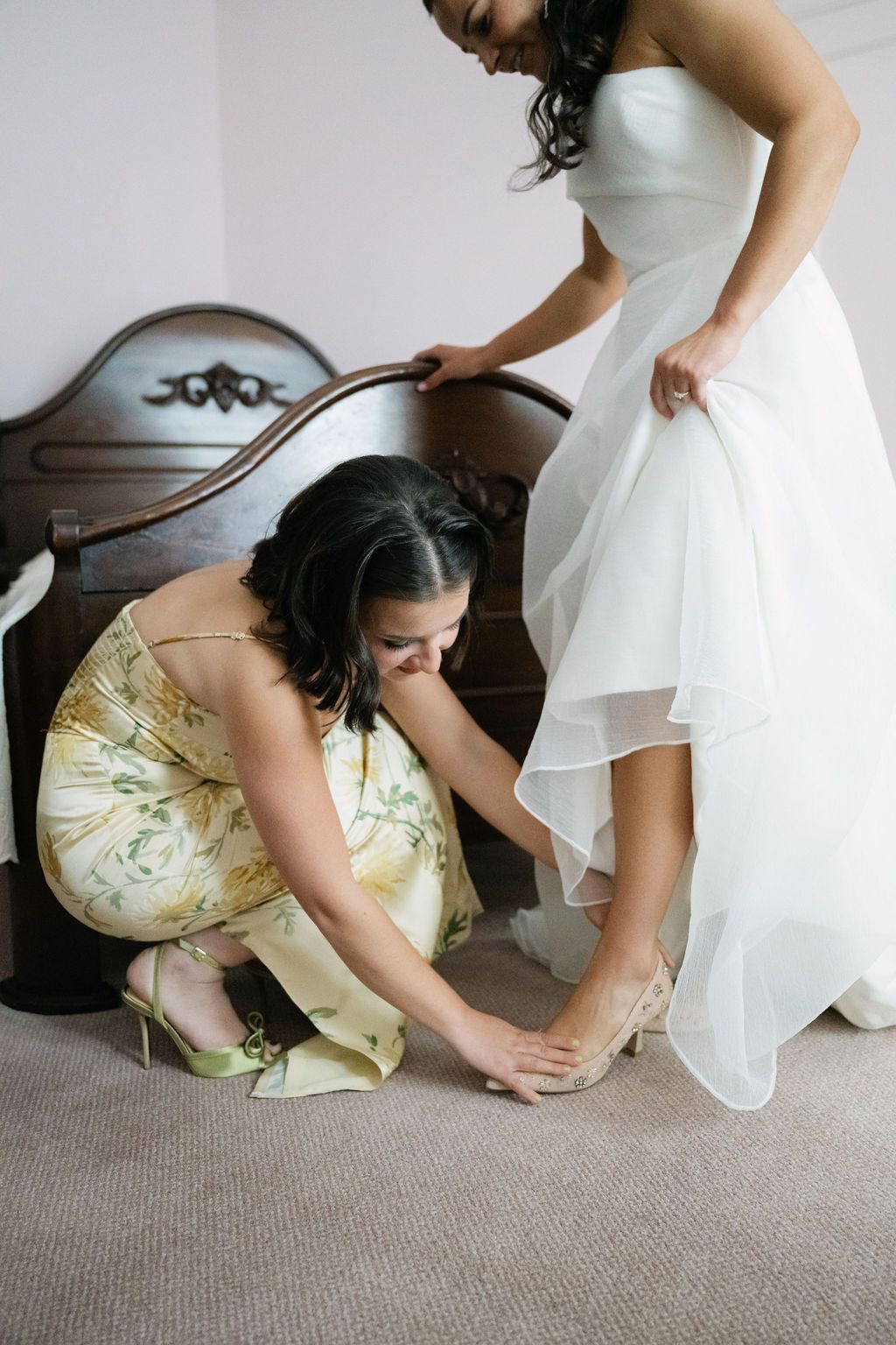 Bridesmaids helping adjust wedding shoes and dress hem on carpeted floor.