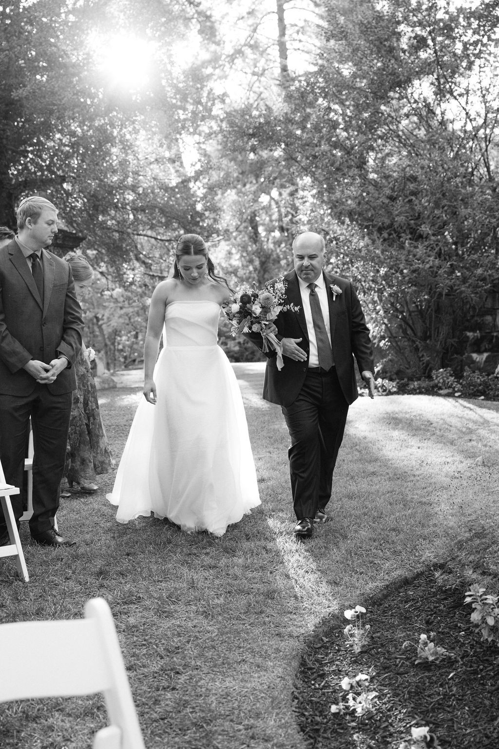 A wedding ceremony processional along a garden path lined with white chairs.