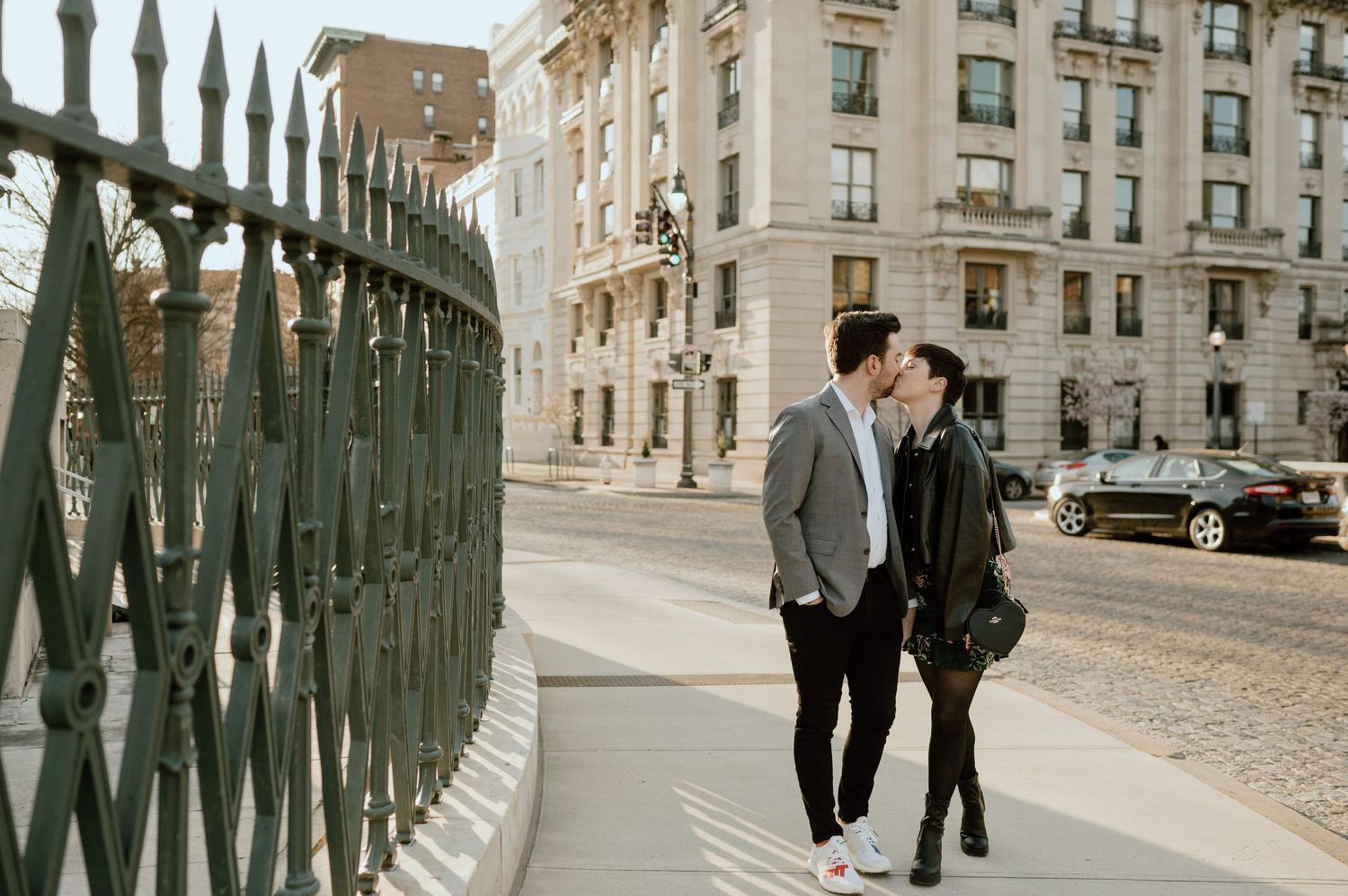 A couple shares a romantic kiss on a sunlit city sidewalk beside ornate green iron fencing with a grand building behind them.
