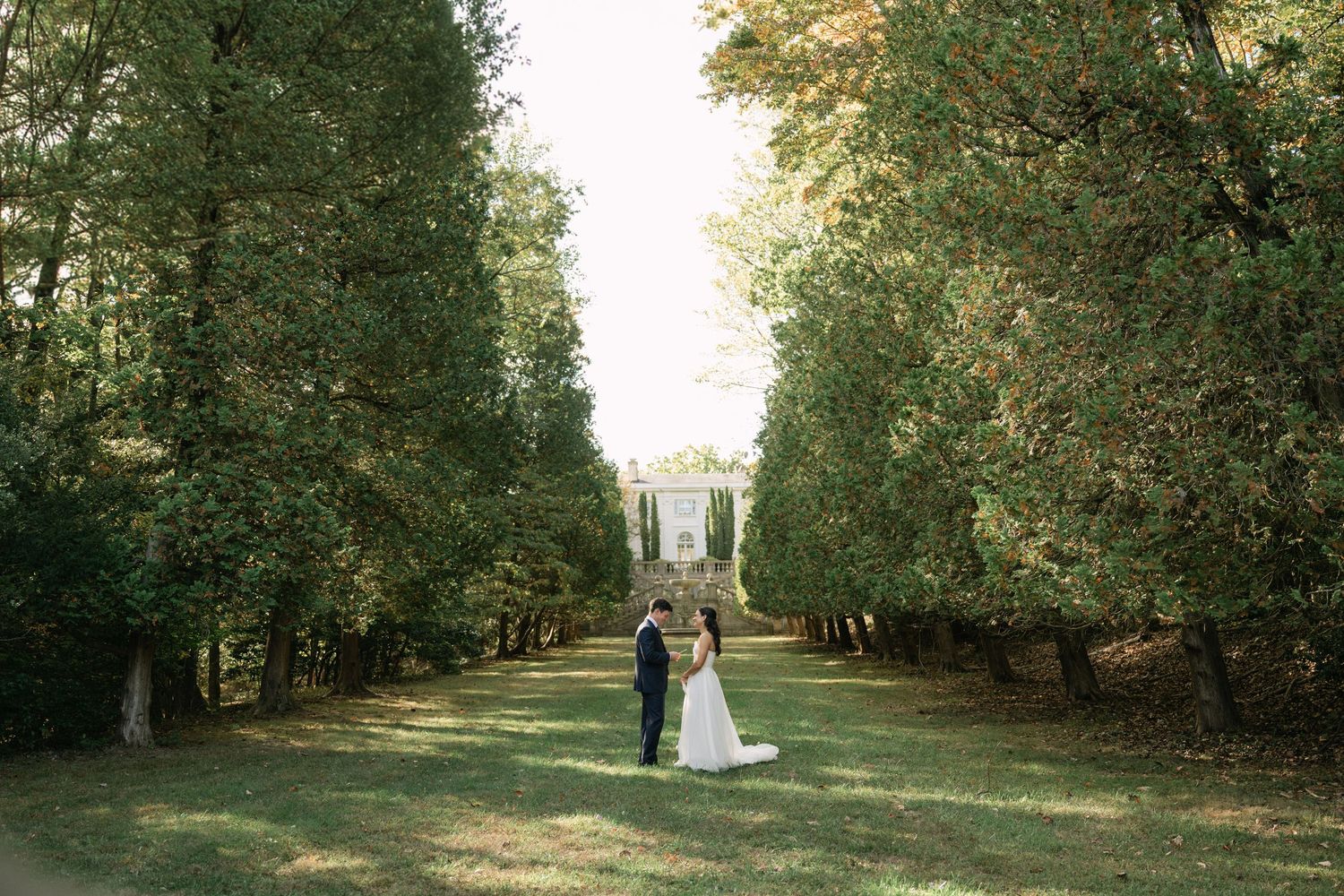 Bride and groom stand together on a lush green tree-lined path leading to an elegant white gazebo.
