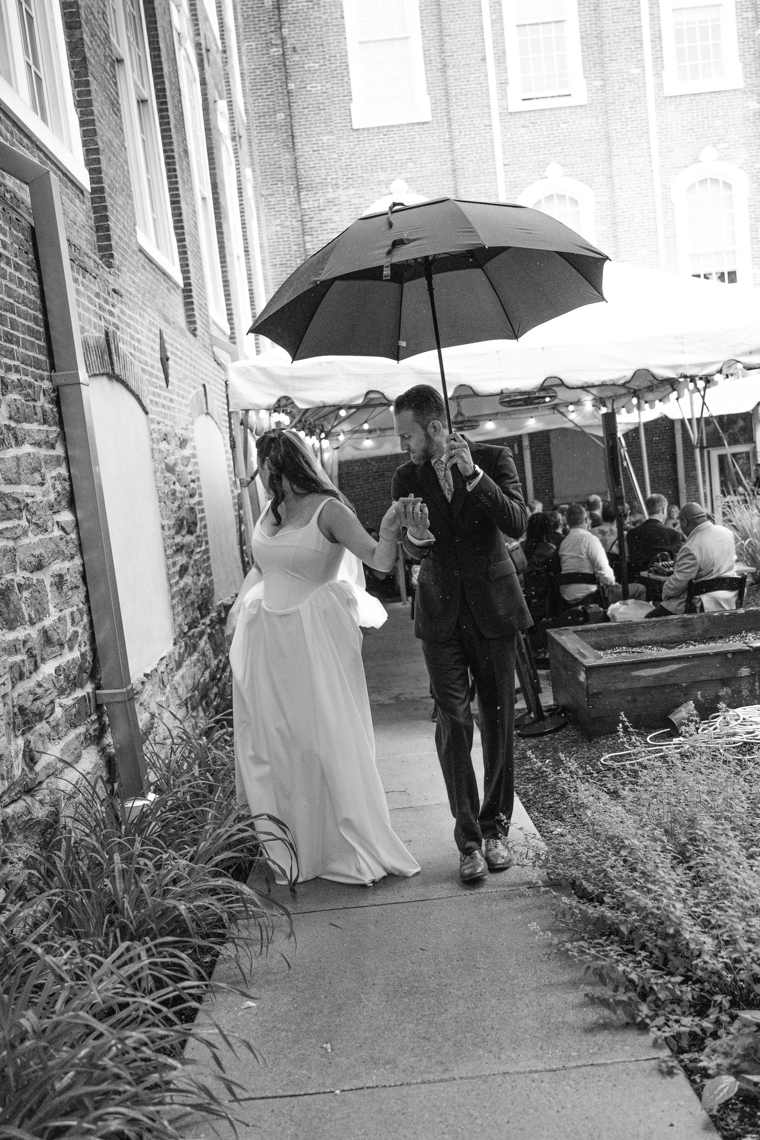 Black and white photo of a bride and groom walking under an umbrella on a rainy wedding day near a brick building.