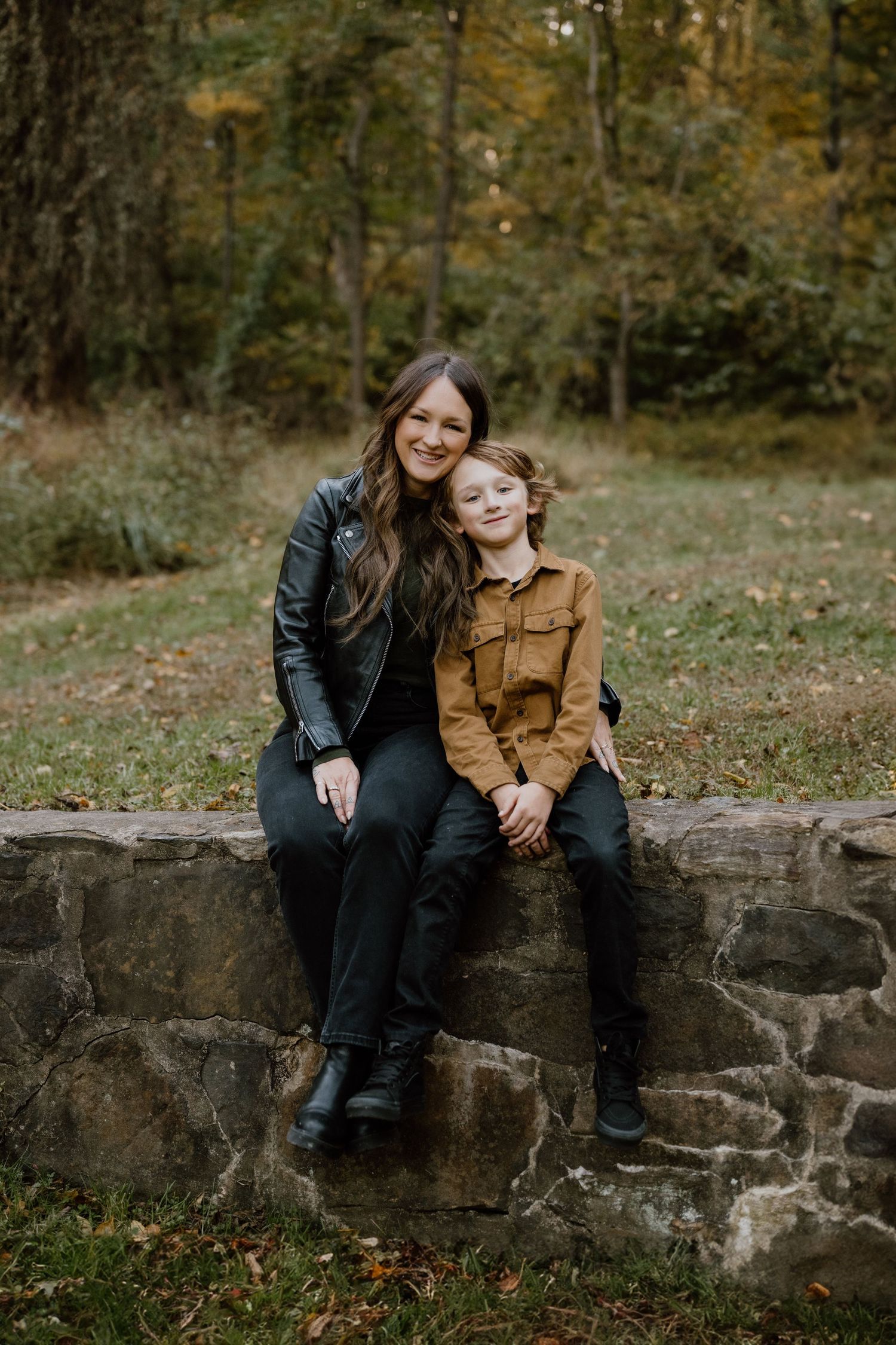 Two people share a relaxed moment sitting on an old stone wall in an autumn forest.