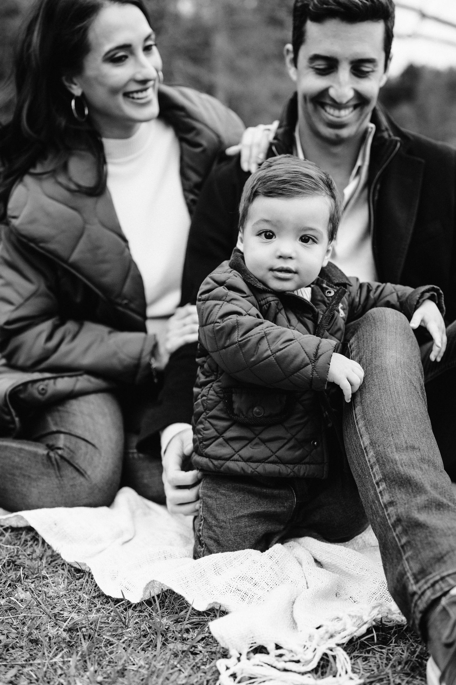 Black and white family photo on blanket outdoors with toddler in quilted jacket.