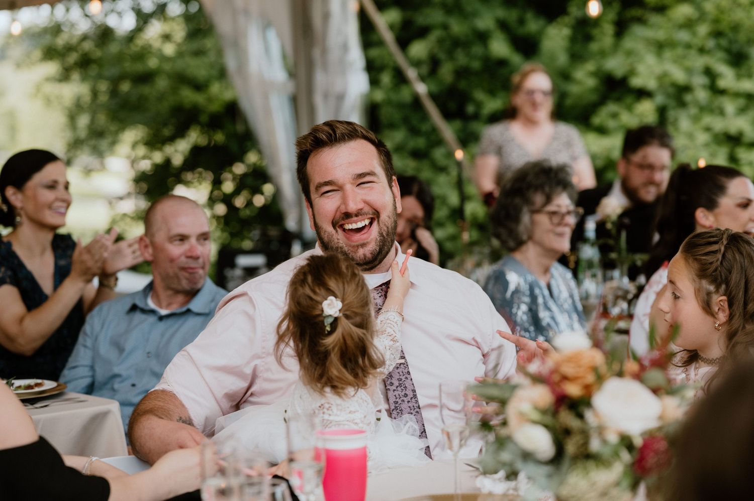 Wedding reception guests share laughter during outdoor celebration under tent with string lights.