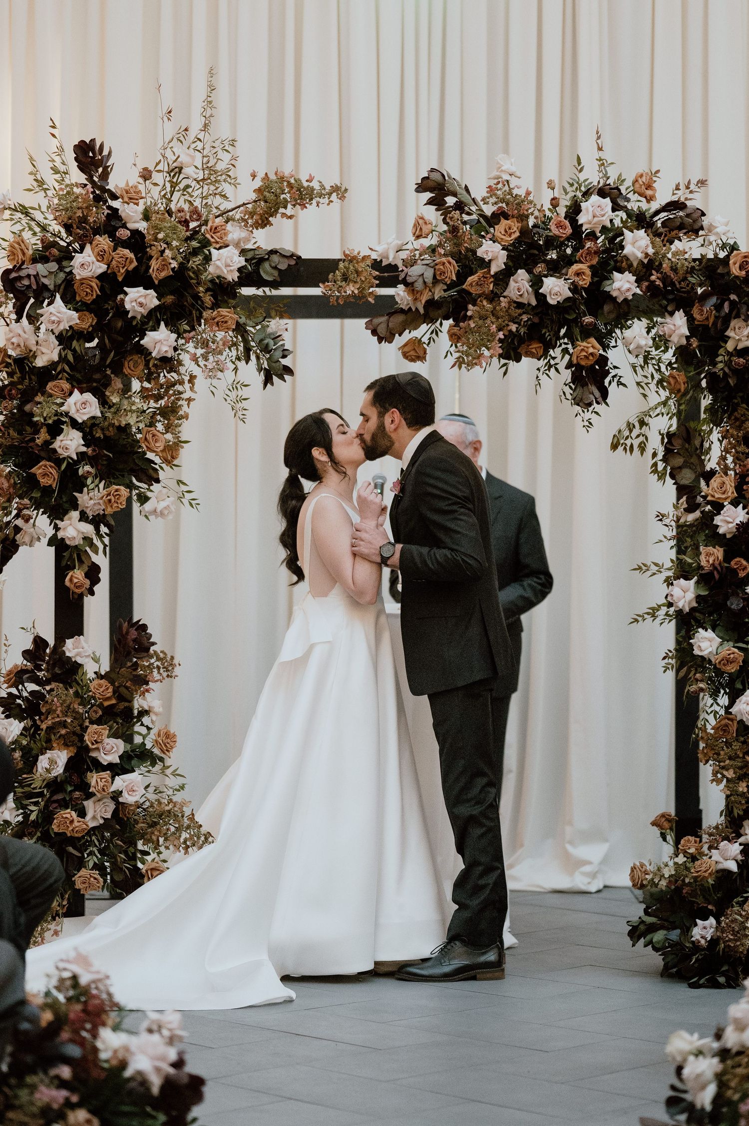 Romantic wedding moment under a lush floral arch with autumn-colored blooms.
