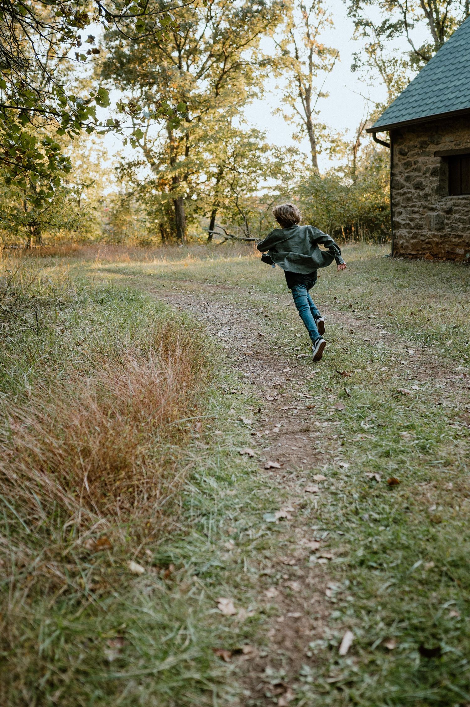 A child runs along a nature trail past a rustic stone building on an autumn afternoon.