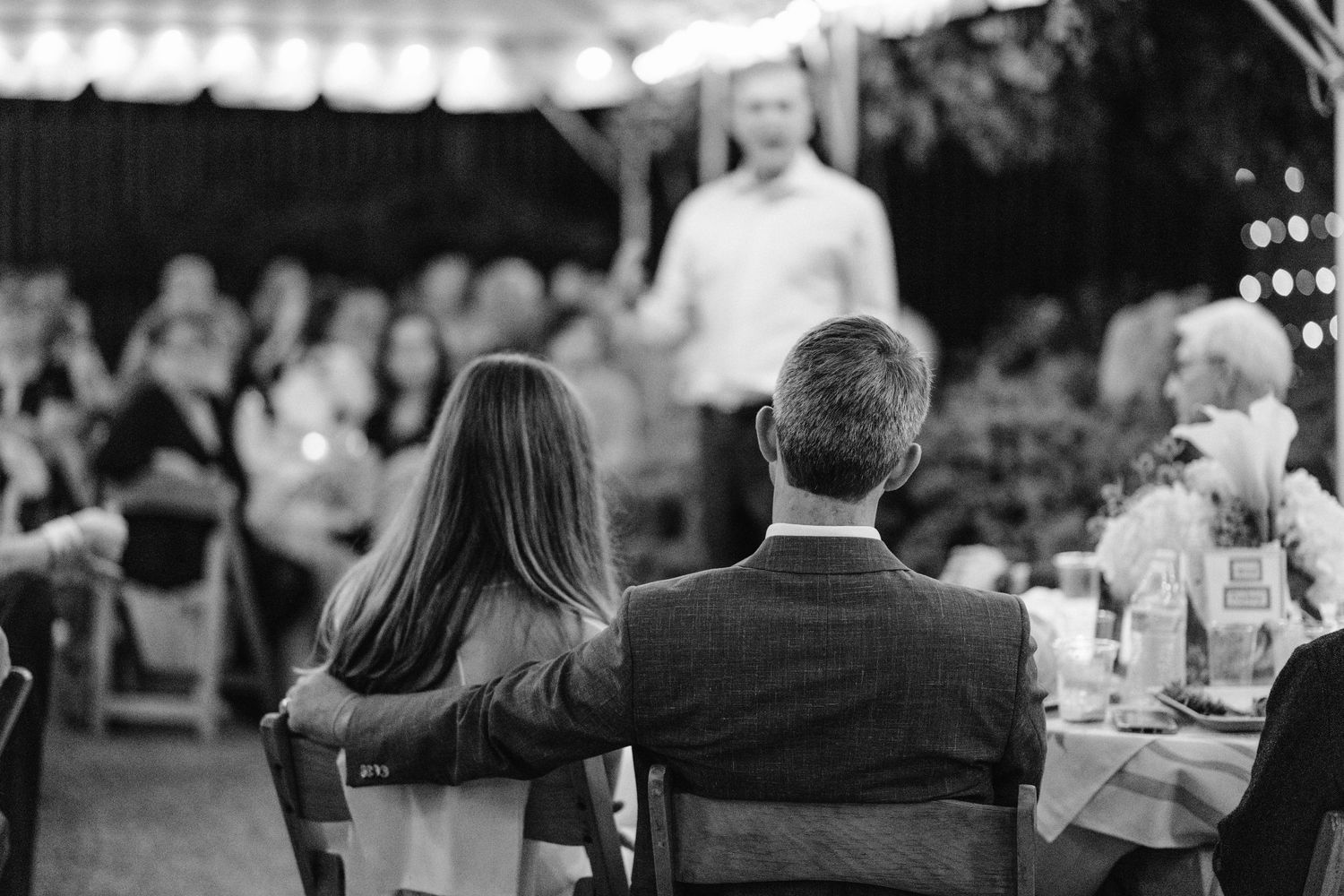 Black and white photo of couple listening to wedding speeches under outdoor tent.