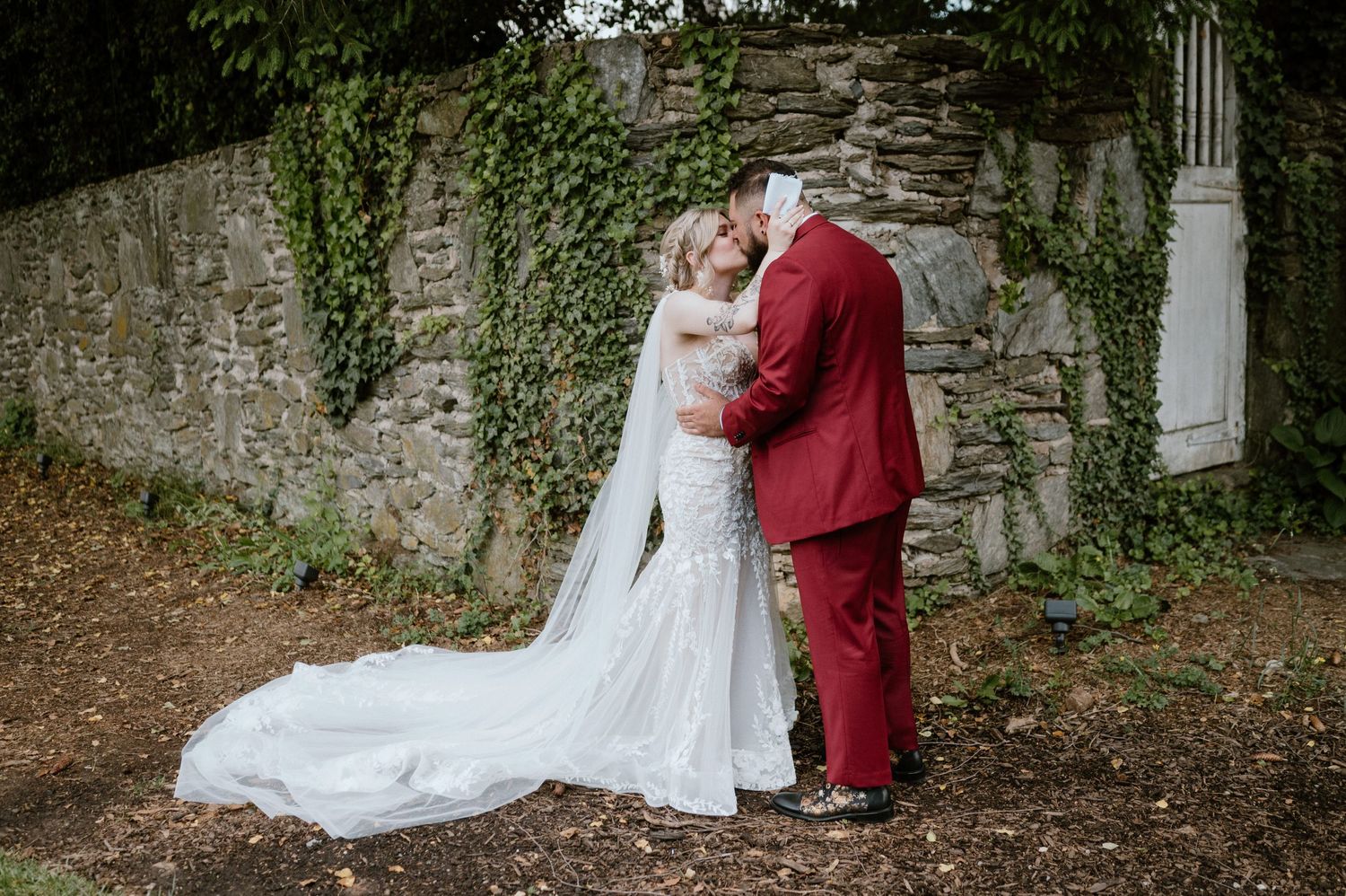 Newlyweds embrace in front of stone wall covered with ivy, bride in white gown and groom in burgundy suit.