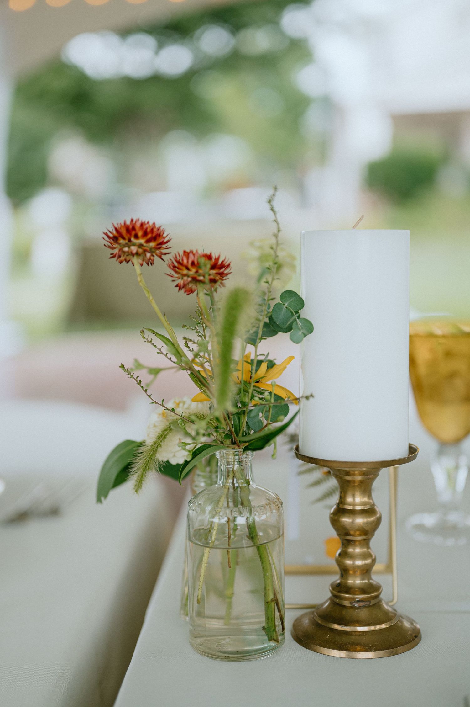 A rustic wedding centerpiece with wildflowers in a glass bottle next to a brass candlestick holder.