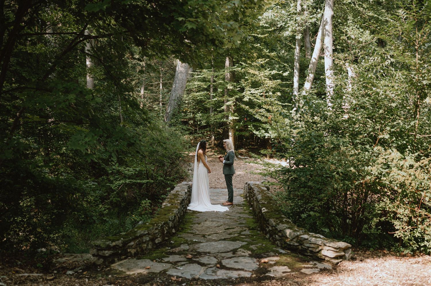 A couple in wedding attire walks along a rustic stone bridge surrounded by forest greenery.