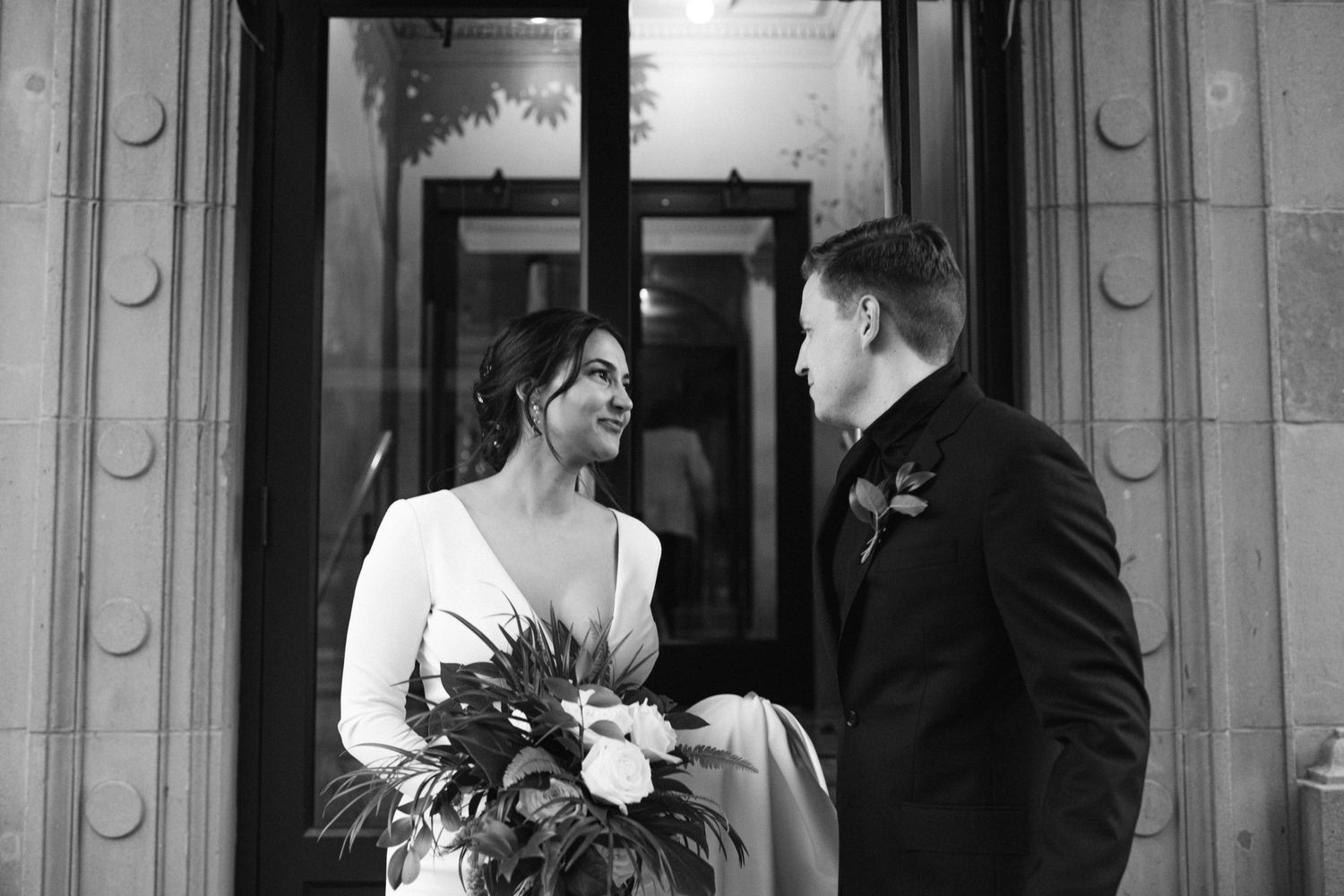 Black and white portrait of a wedding couple with bouquet in front of ornate doorway.