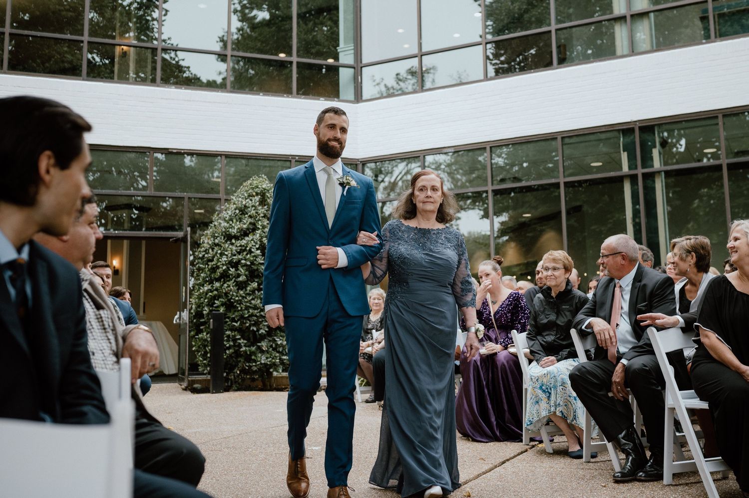 Wedding processional moment outside modern glass building with guests watching from white chairs.
