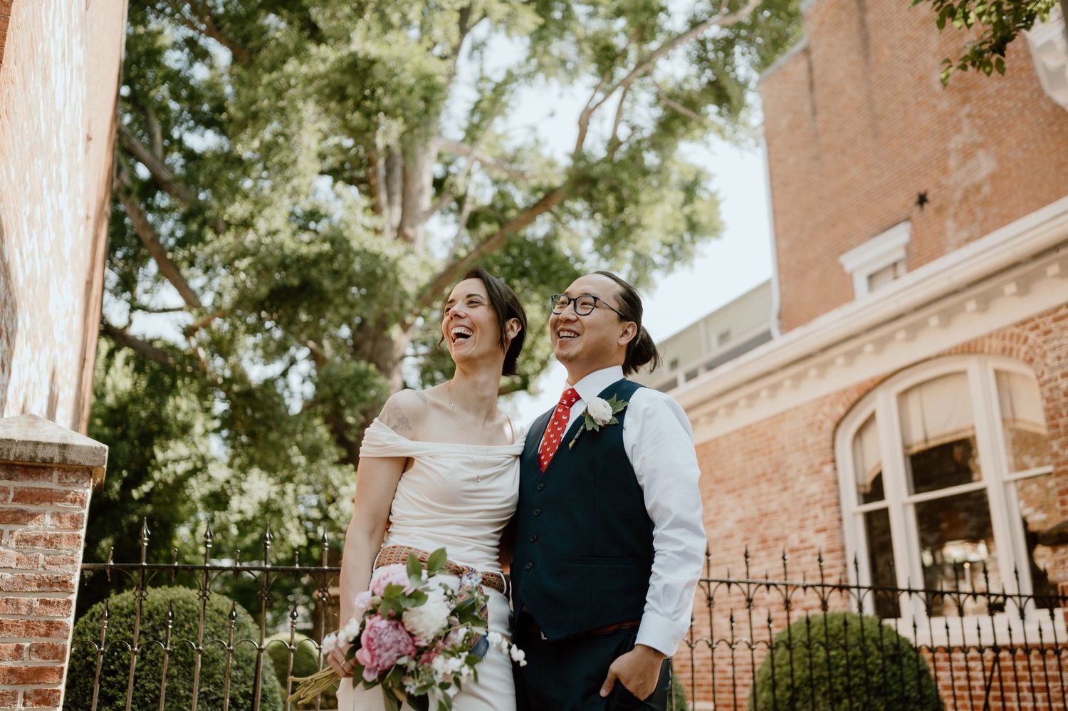 A couple shares a joyful moment together in wedding attire against a historic brick building.