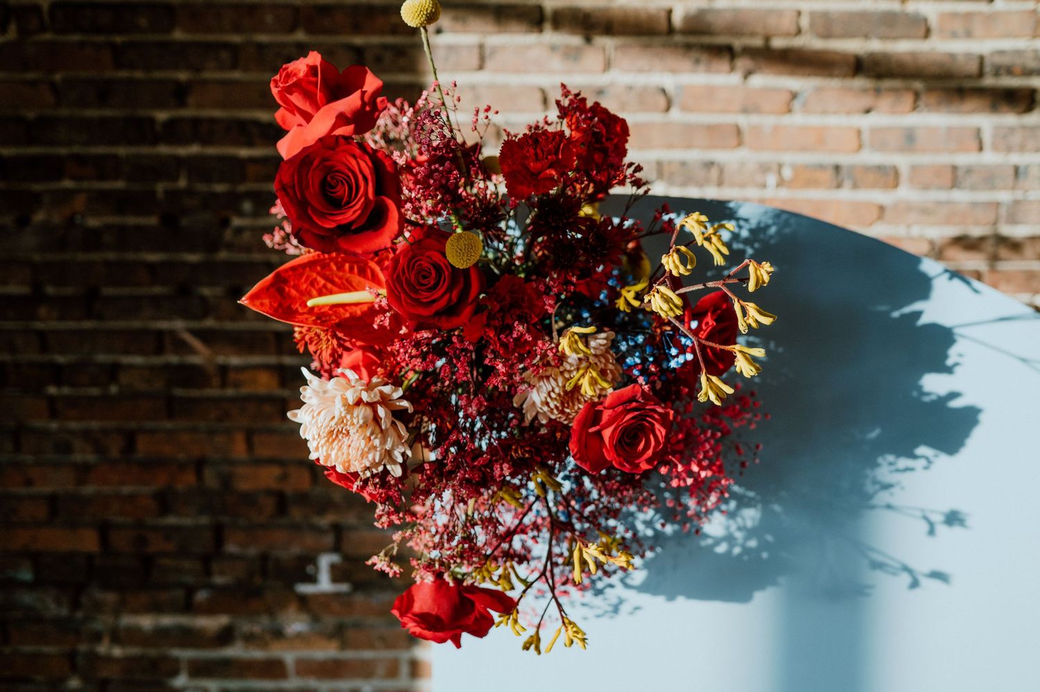 Vibrant red and burgundy floral arrangement casting dramatic shadows against brick wall.