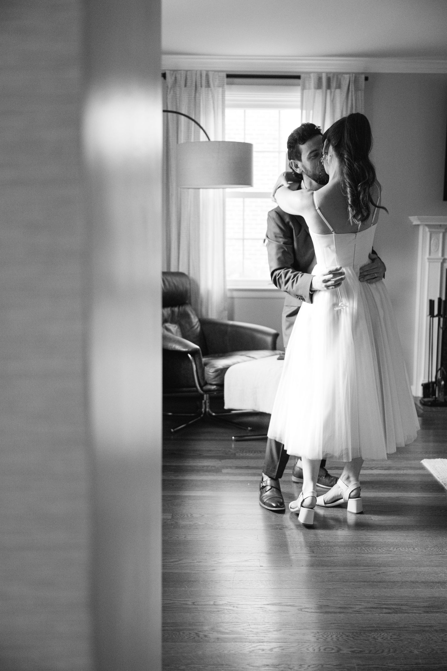 A romantic black and white photo of a couple dancing in a sun-lit room with flowing curtains and hardwood floors.
