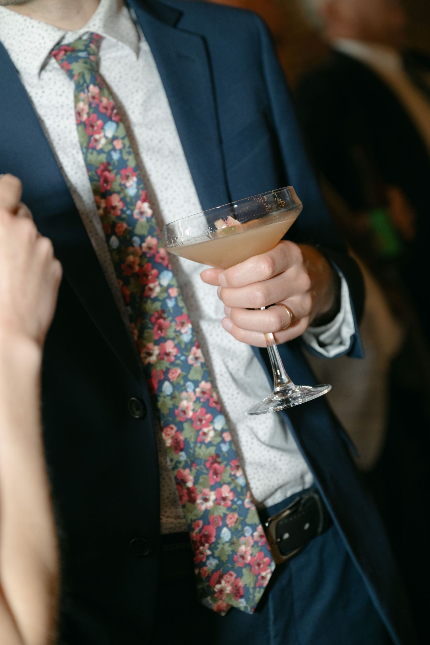 Two hands holding wine glasses with a navy suit and floral tie visible in formal attire.