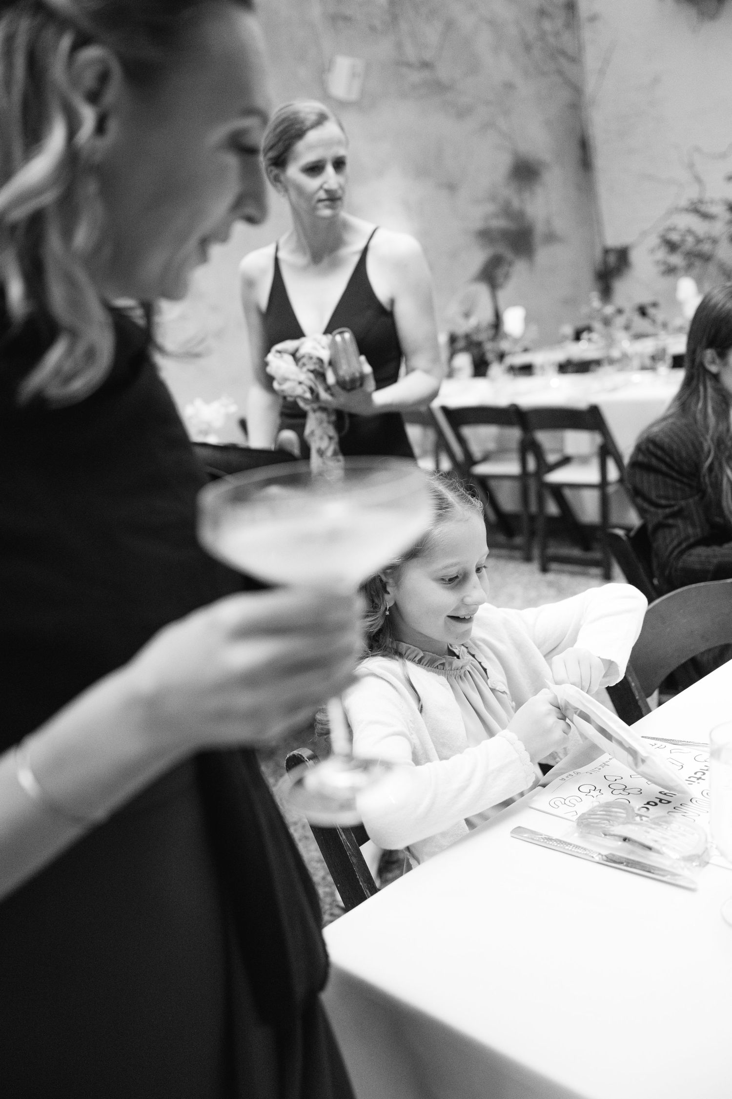 Black and white photo of dining guests at a reception with table settings and folding chairs in background.