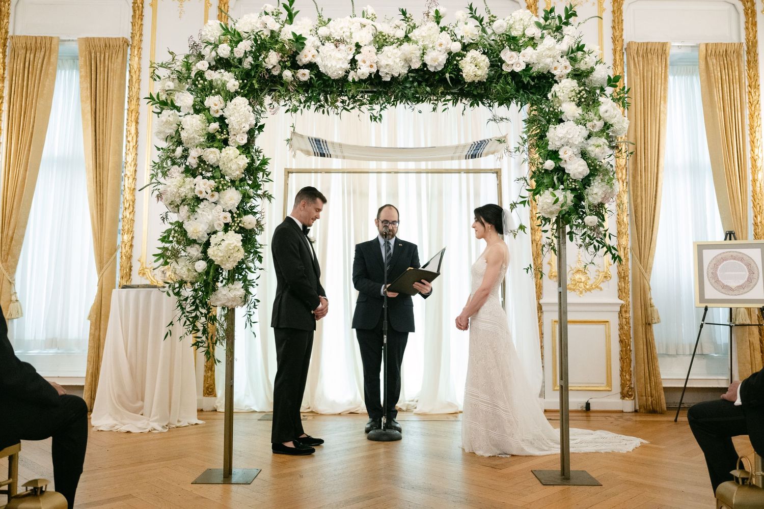 Wedding ceremony setup with a white floral arch and gold details in an elegant ballroom.